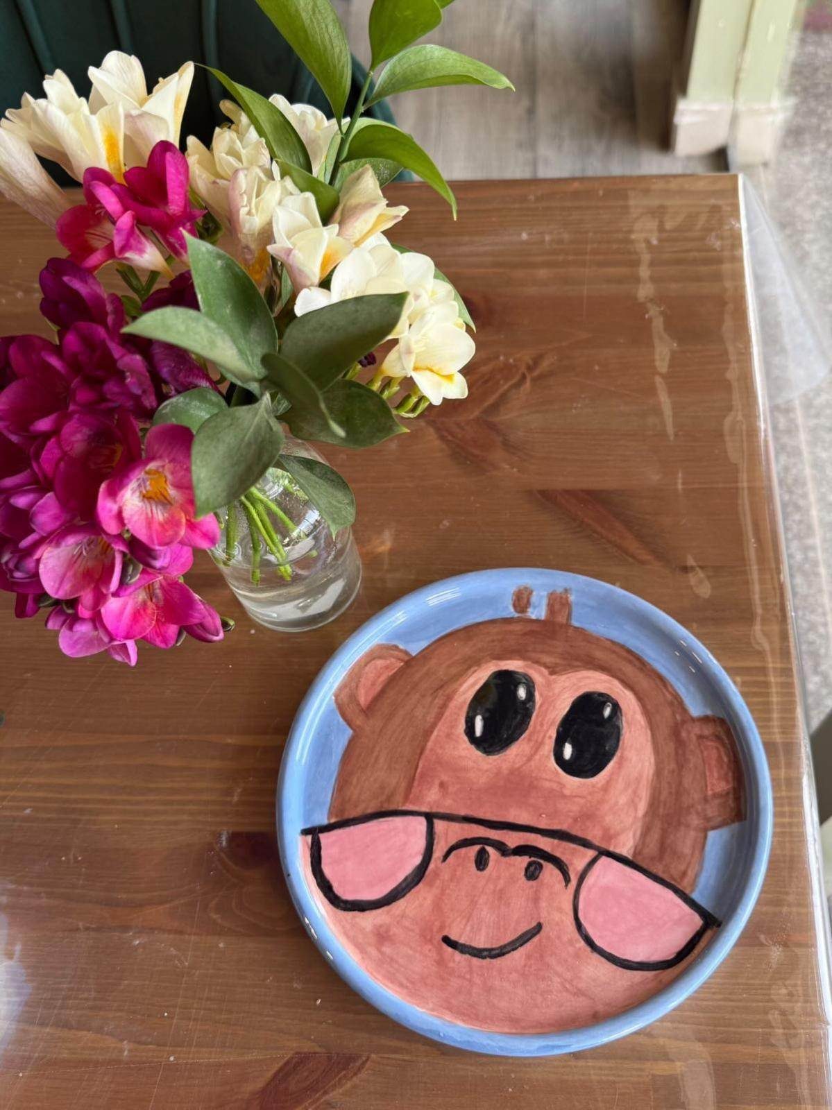 A hand-painted ceramic plate with a cartoon monkey face on a wooden table at Art Bodega, beside a jar of pink and white flowers