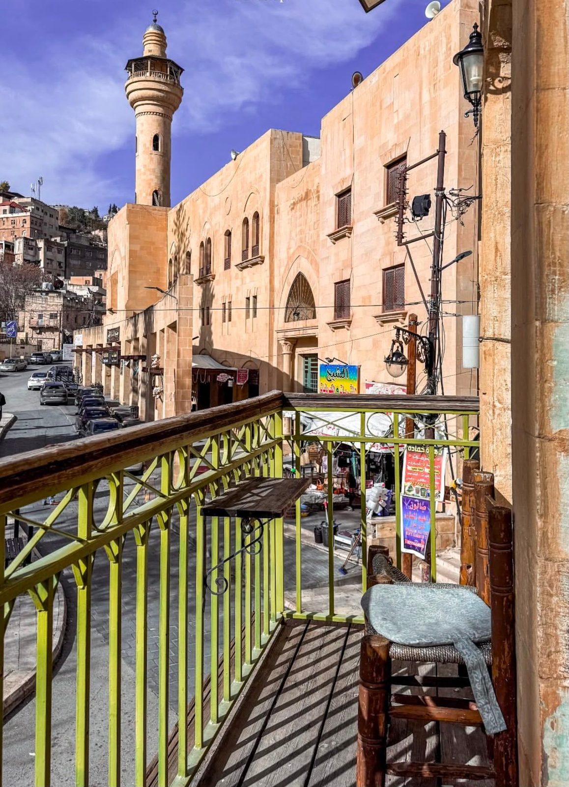 A wooden stool on a green-railed balcony in Jabal Al Weibdeh, looking across the street to a stone mosque and its minaret